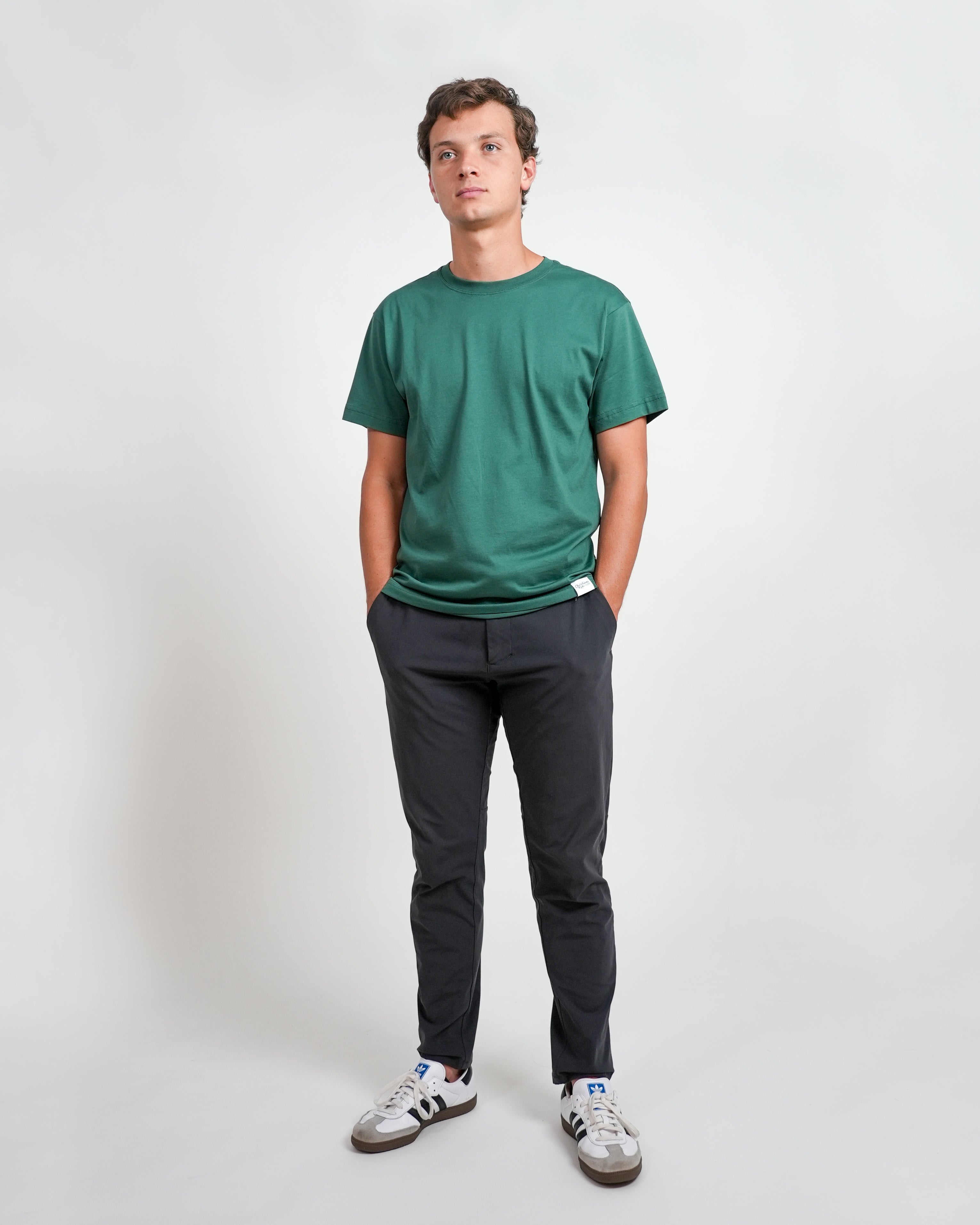 Smiling male model wearing a green t-shirt with brand logo and black pants standing against a white background.