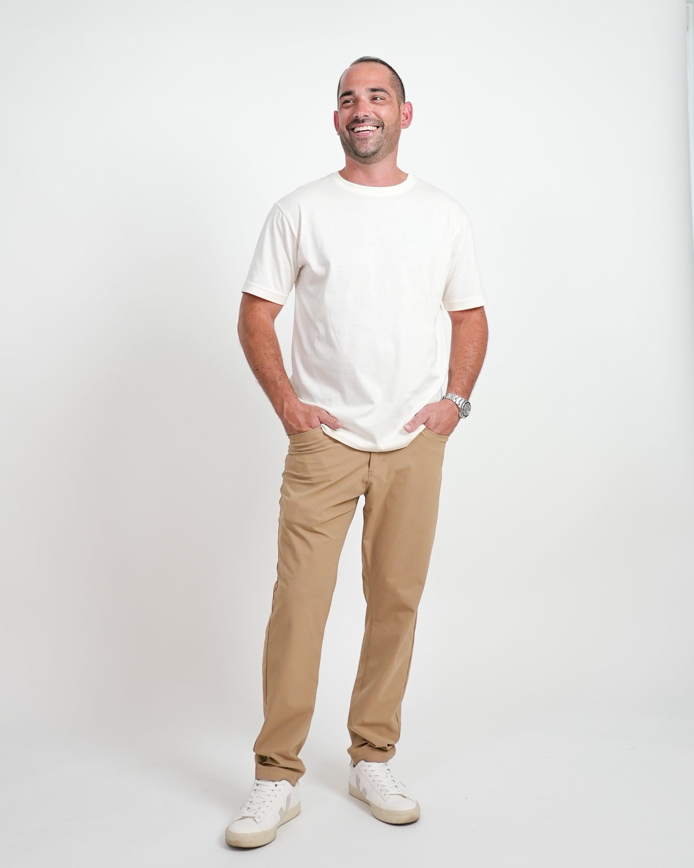 Smiling male model wearing a cream t-shirt and beige pants, standing in front of a white background.