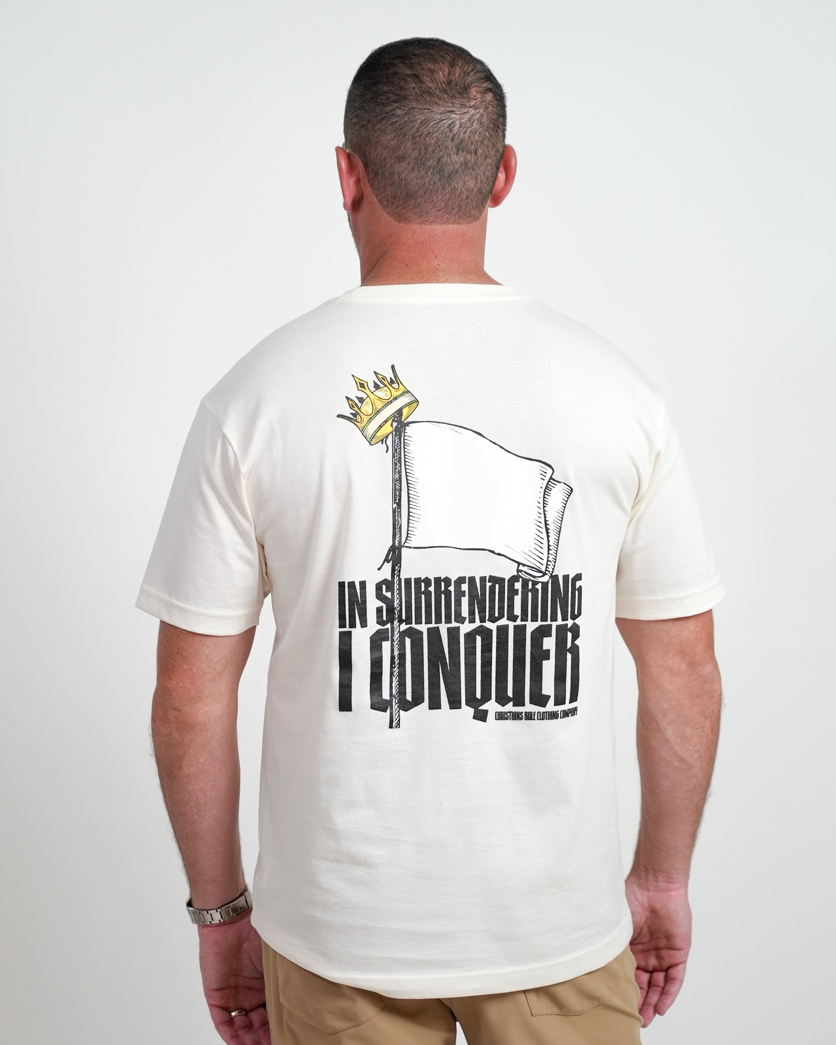 Male model wearing a cream Christian t-shirt with a flag graphic design and black text saying "In surrendering I conquer" and "Christians Rule Clothing Company." Person is standing in front of a white background.