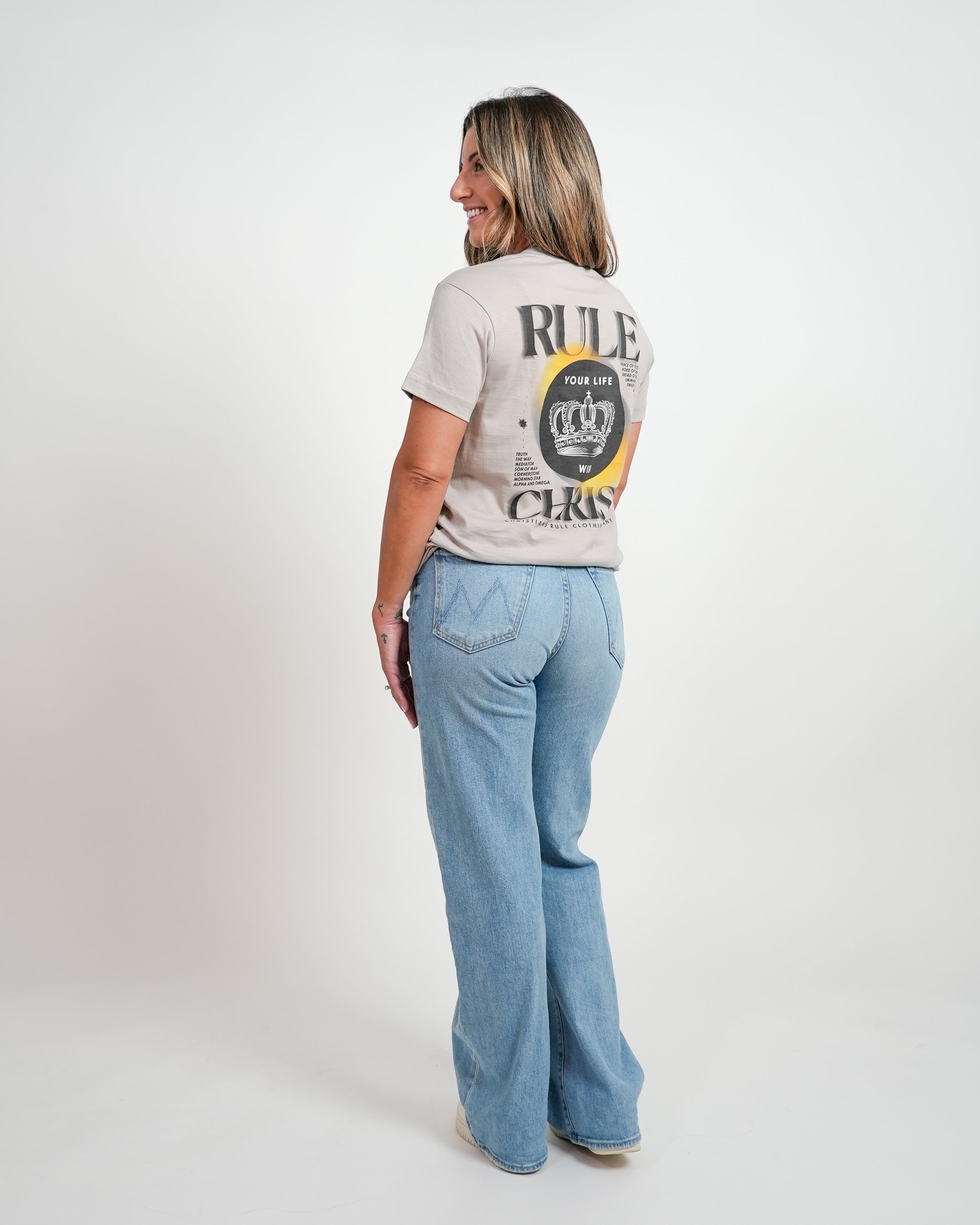 Smiling female model wearing gray Christian t-shirt with "Rule Your Life with Christ" graphic and jeans, standing against a white background.