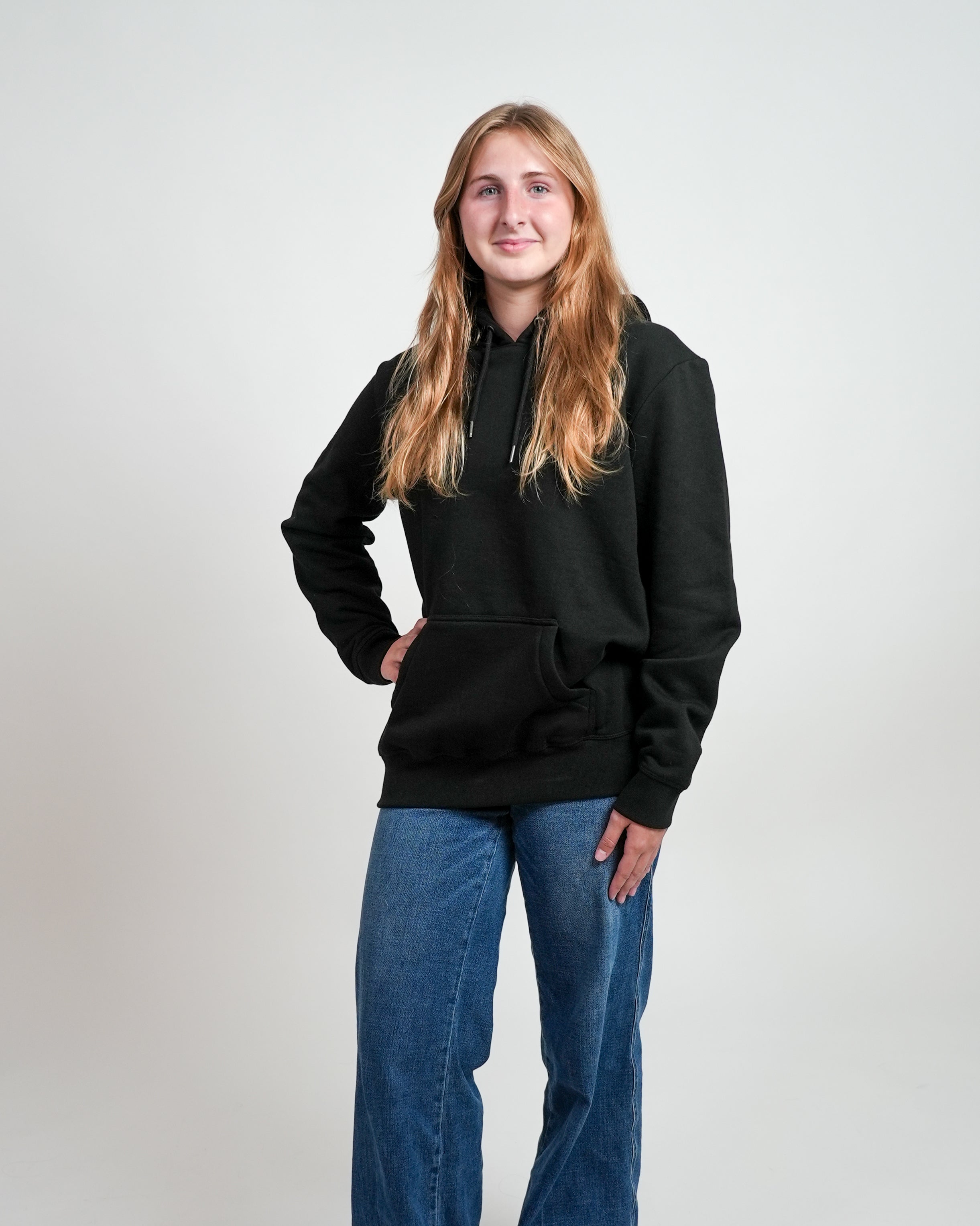 Smiling female model wearing a black hoodie and jeans, standing against a white background.