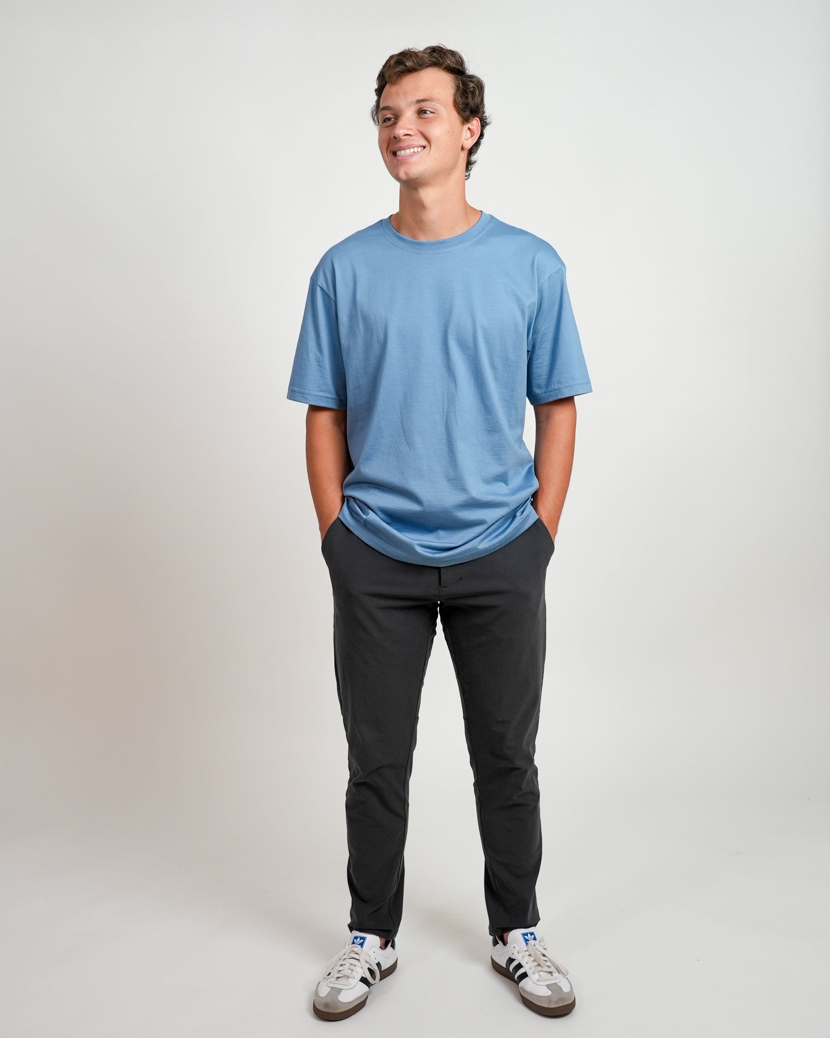Smiling male model wearing blue t-shirt and black pants standing against a white background.