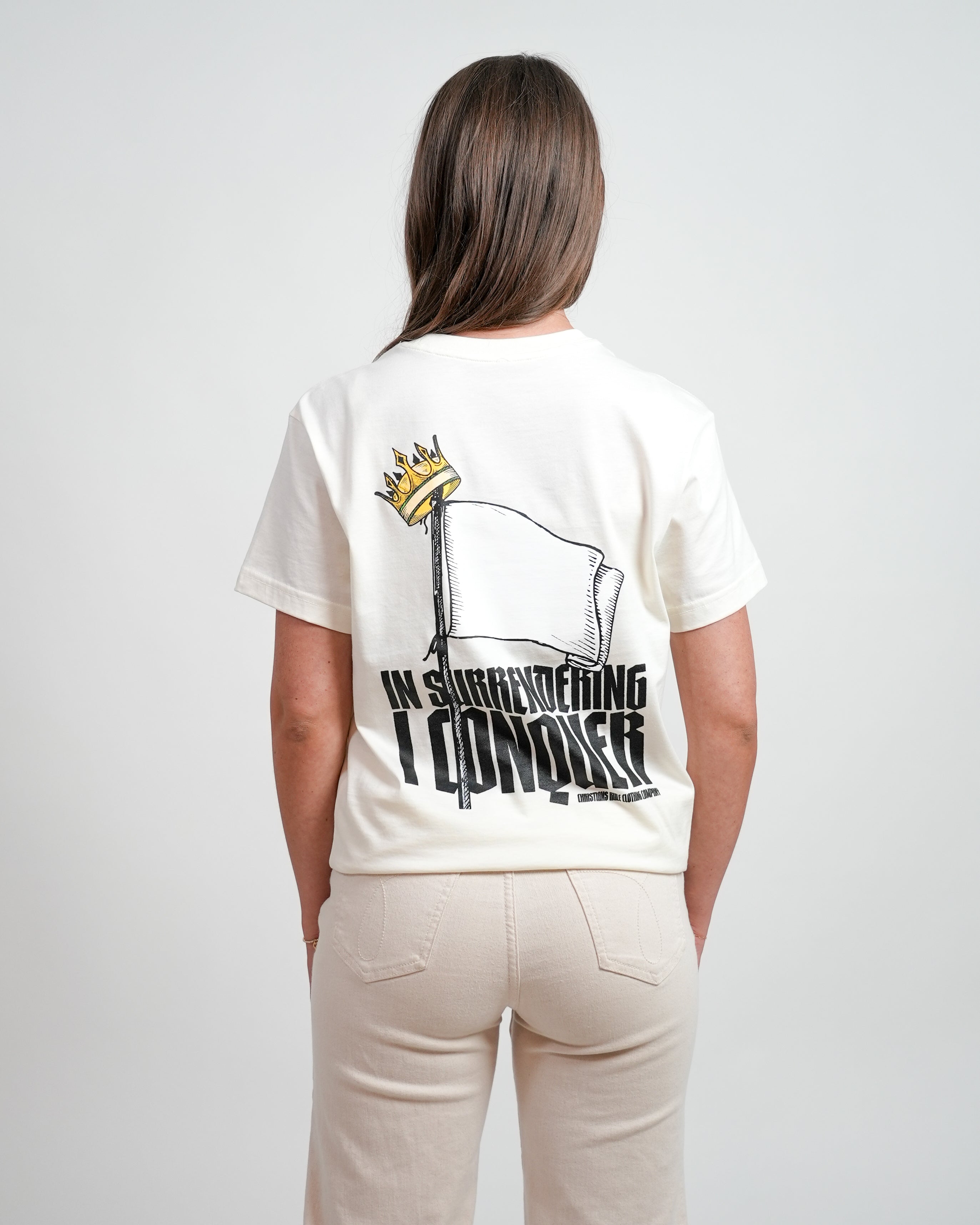 Female model wearing a cream Christian t-shirt with a flag graphic design and black text saying 'In surrendering I conquer' and 'Christians Rule Clothing Company.' Person is standing in front of a white background.