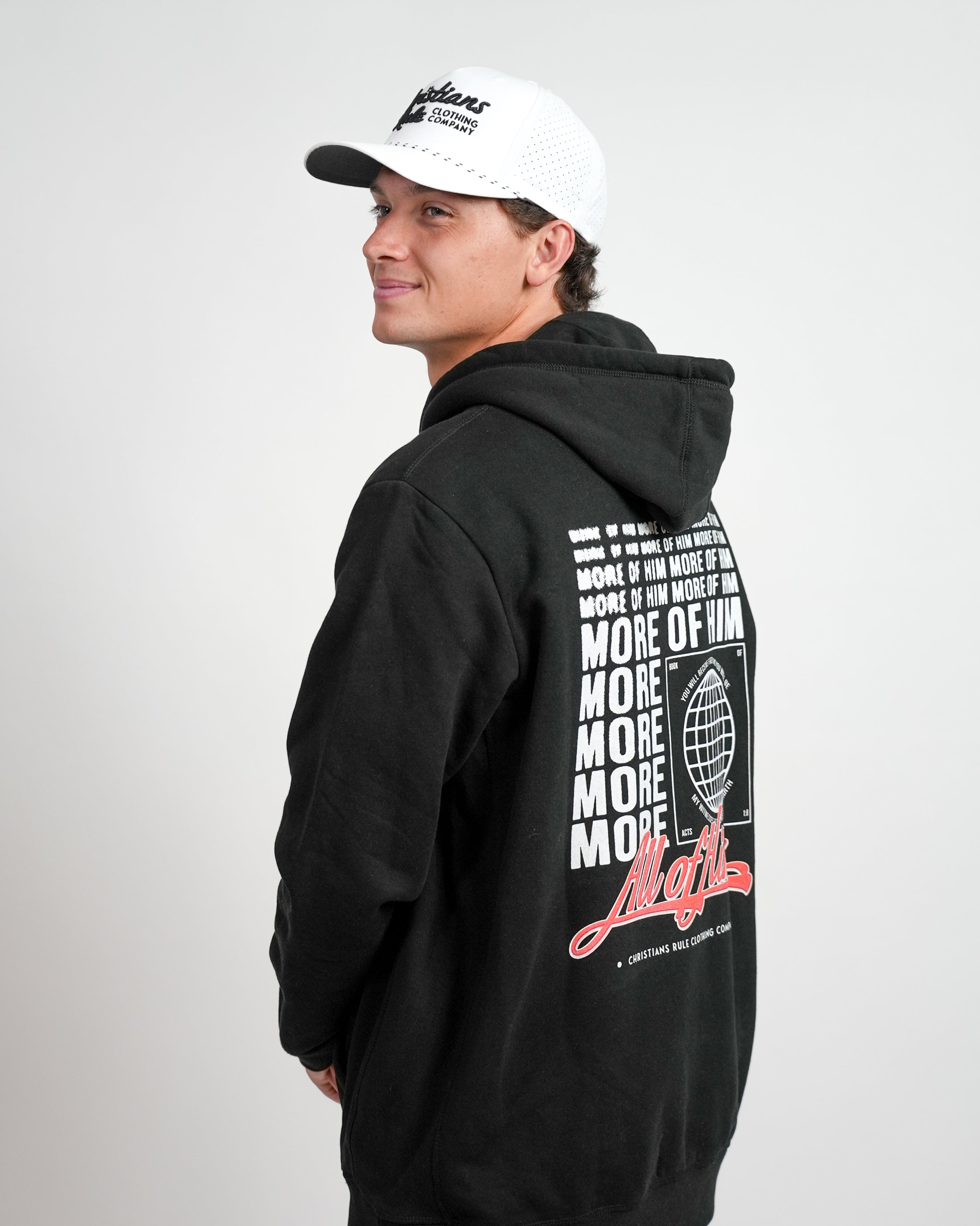 Male model wearing a white Christian golf rope cap with 'Christians Rule Clothing Company' embroidered text and black hoodie with 'All of Him' script graphic, standing against a white background.