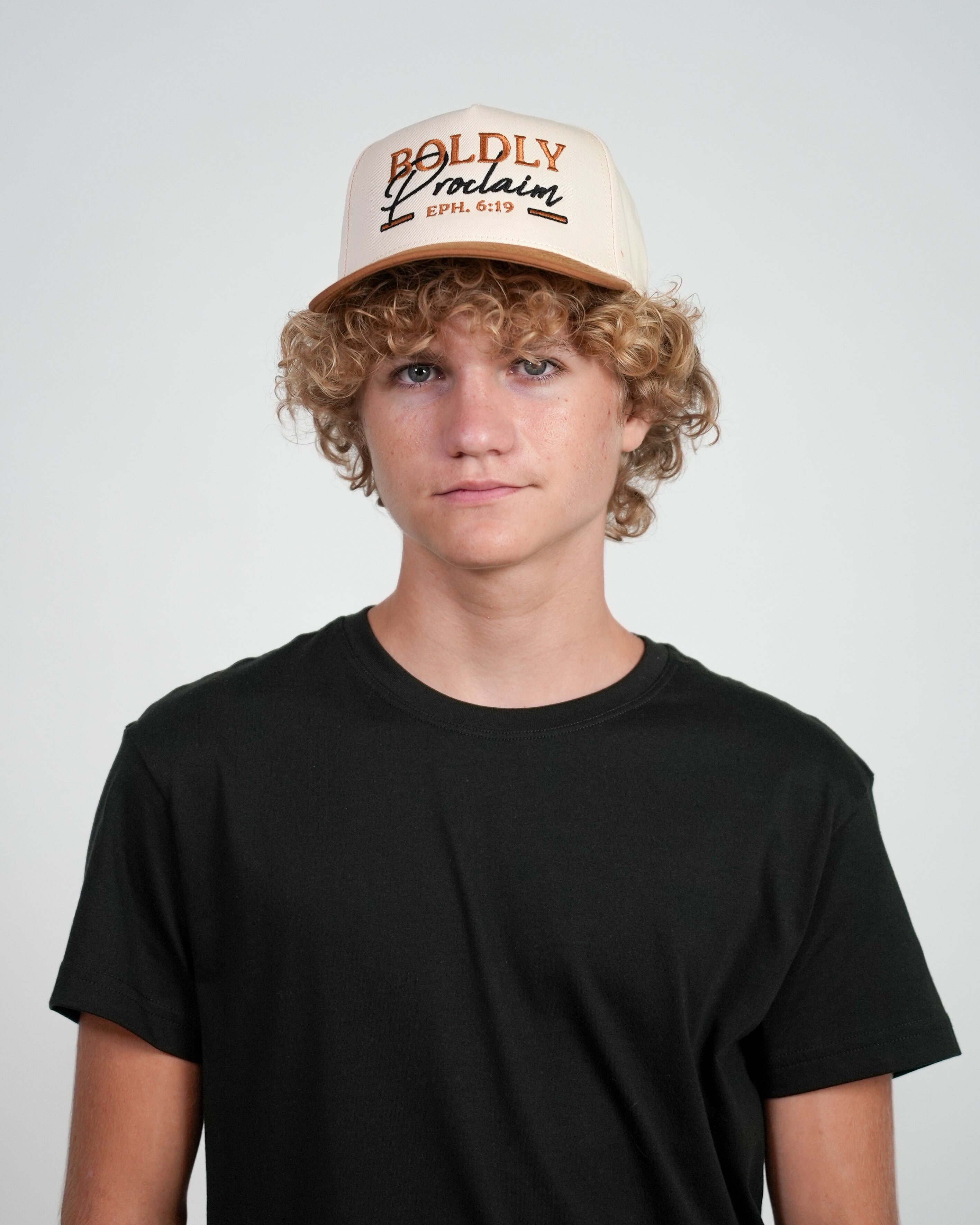 Male model wearing a cream and bronze Christian cap with embroidered text that says 'Boldly Proclaim', standing against a white background.