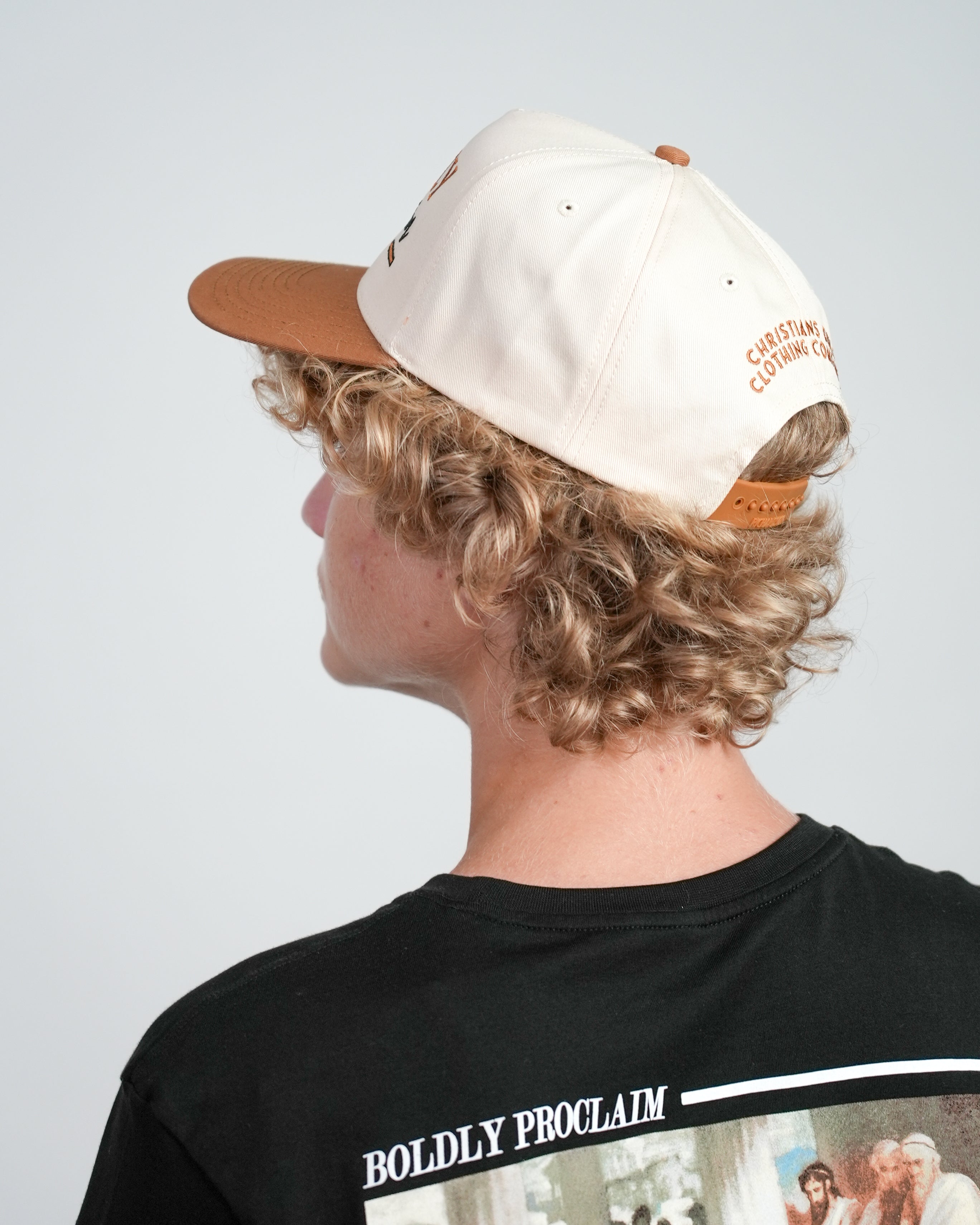 Side view of a male model wearing a cream and bronze Christian cap with embroidered text, against a white background.