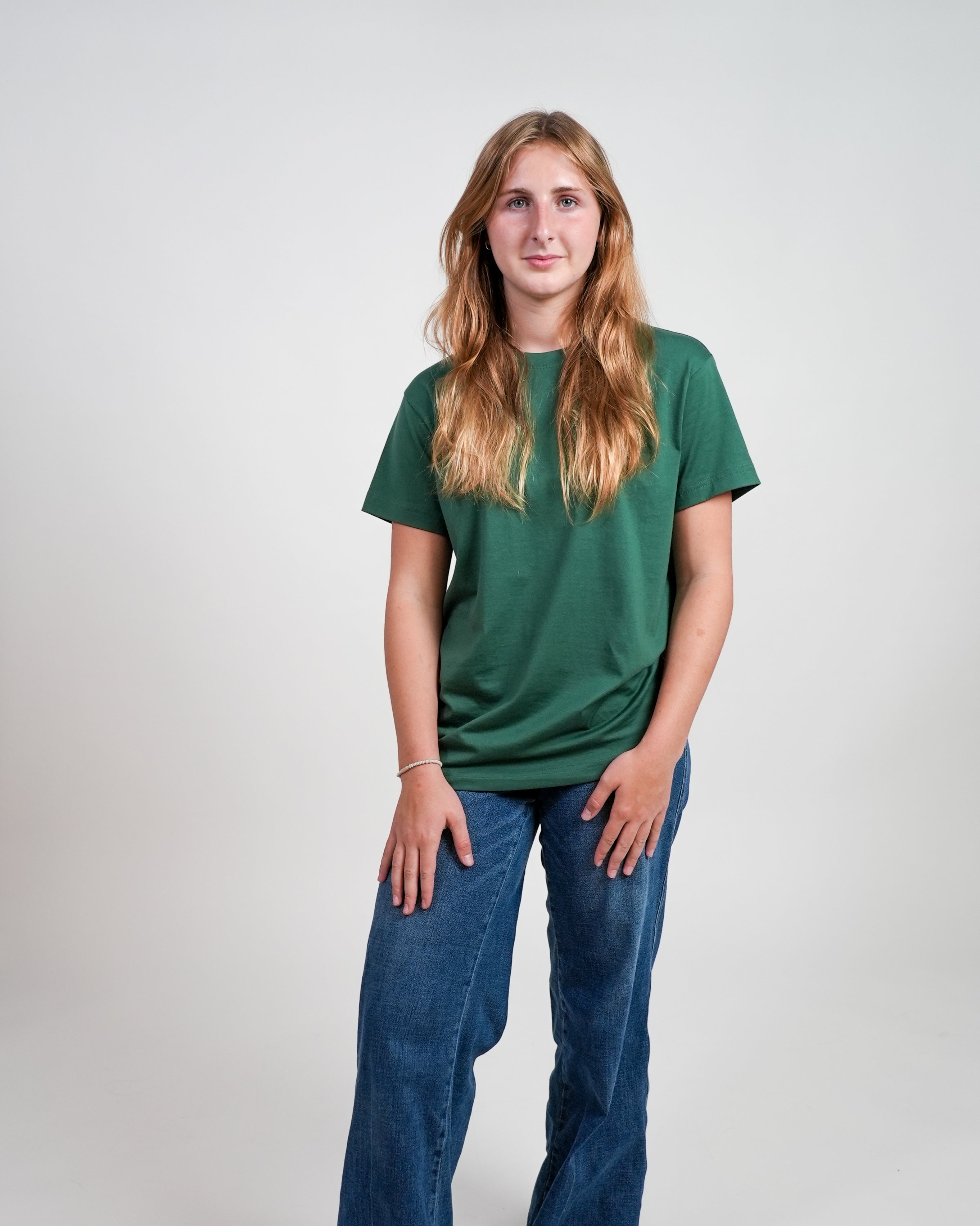 Smiling female model wearing a green t-shirt and jeans standing against a white background.