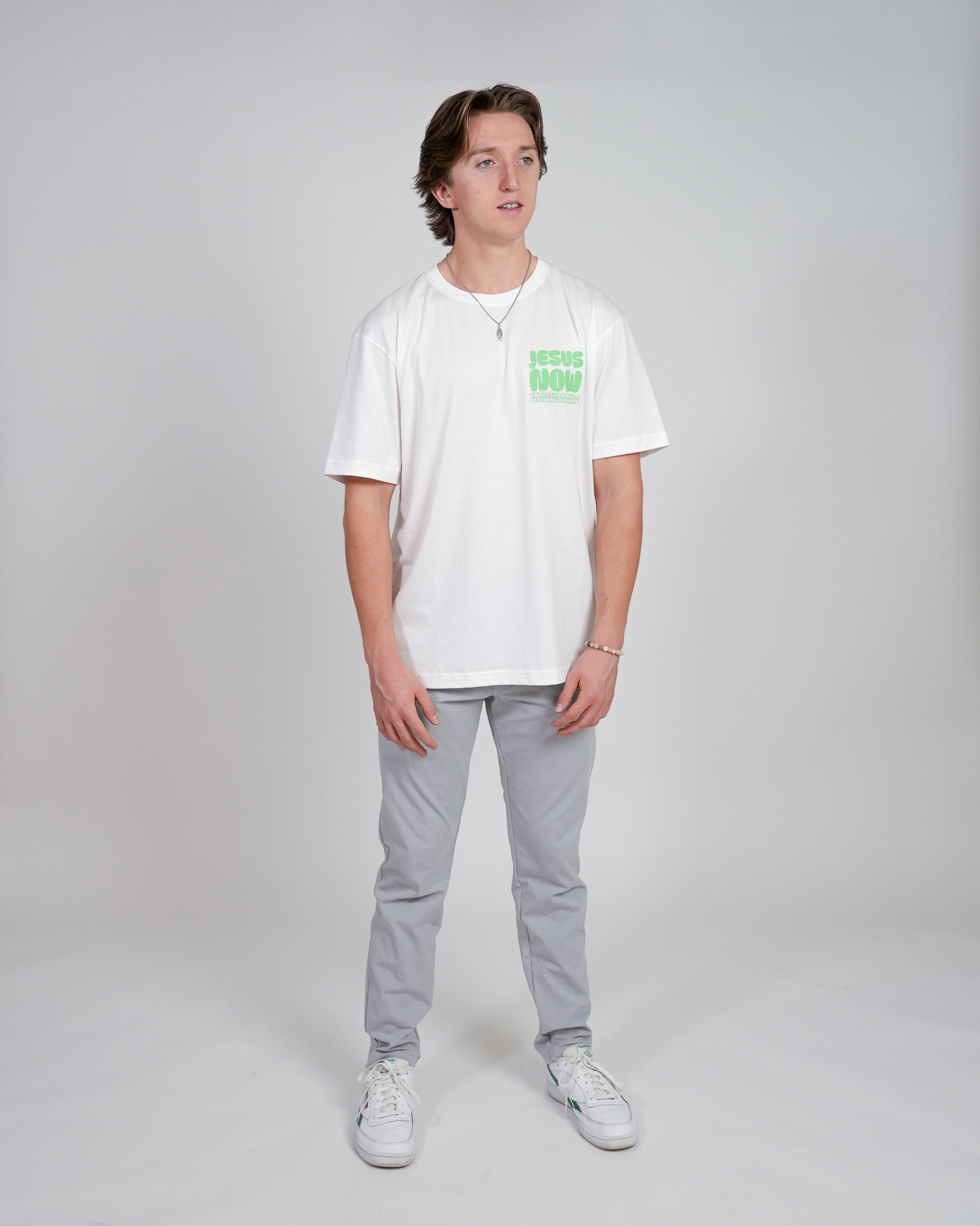Smiling male model wearing a white Christian t-shirt with 'Jesus Now' graphic design and gray pants, standing against a white background.